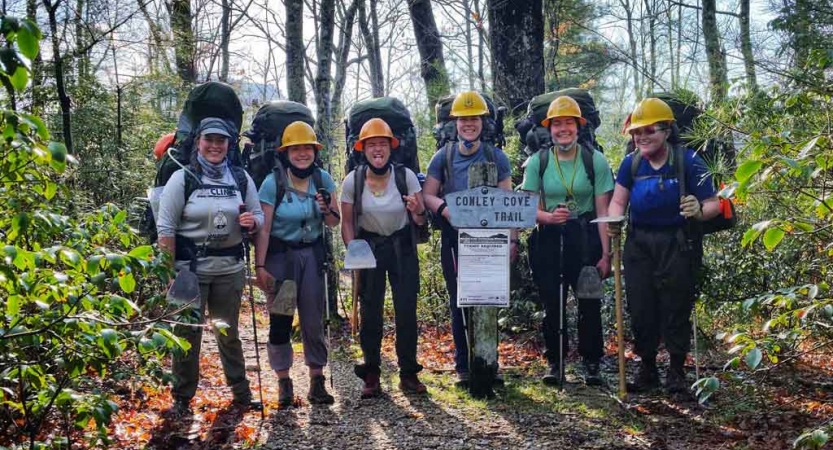A group of people wearing helmets and backpacks pose for a photo near a trail sign. 